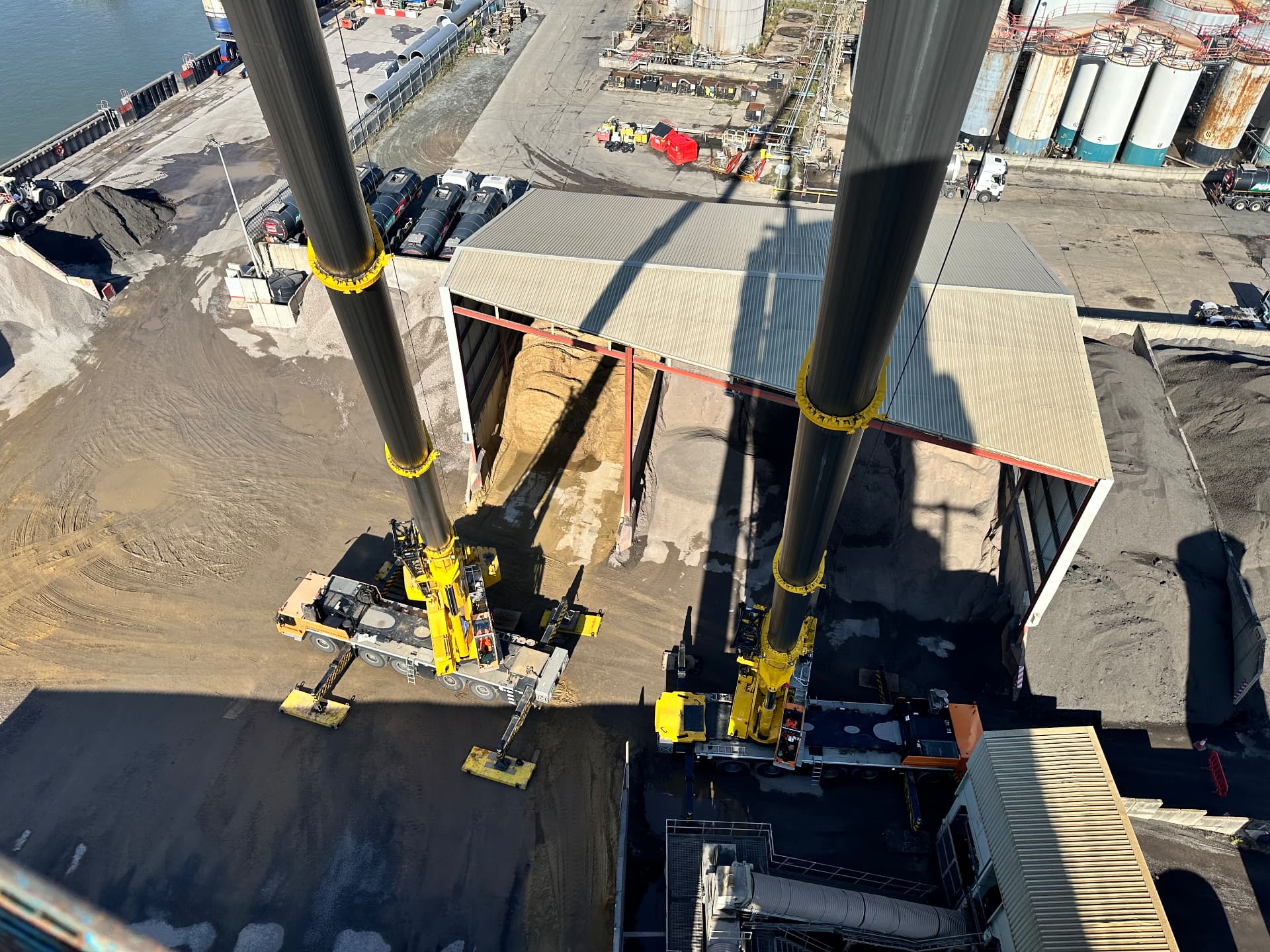 Aerial view of a tandem lift raising two large steel tubes at a dock